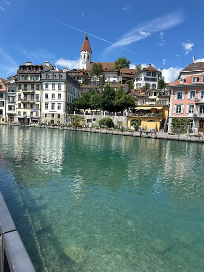 The image depicts a sunny day in the city of Thun, Switzerland, with a clear blue sky dotted with white clouds and a contrail from an airplane. The foreground features a body of remarkably clear turquoise water, revealing the rocky riverbed below. A dark railing runs along the bottom left of the frame.

Along the riverbank, a row of multi-story buildings lines the waterfront. The architecture is varied, with some buildings featuring light-colored facades, balconies, and numerous windows, while others are painted in warmer tones like yellow and peach, some with red-tiled roofs. Many of these buildings have awnings over ground-level windows, suggesting shops or cafes. A prominent white tower with a red spire and a clock face rises above the other structures, indicating a church or historical landmark. Trees and greenery are interspersed among the buildings and along the riverbank.

Several people are visible along the riverside promenade. Three individuals are standing near the water's edge, seemingly engaged in conversation or observing the river. One person is wearing a red shirt, another a blue shirt, and the third a lighter-colored top. Further down the bank, a few more figures can be seen in front of the buildings, some potentially patrons of a cafe with outdoor seating under a yellow awning. Bicycles are also parked along the sidewalk in front of one of the buildings. The reflections of the buildings and the sky are visible on the rippling surface of the water.