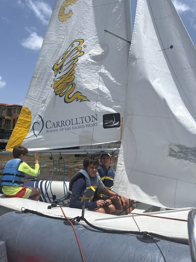 Three boys are in a sailboat on a body of water, with buildings in the background. The sailboat has a white sail with a large yellow and blue logo that reads "CARROLLTON SCHOOL OF THE SACRED HEART". The boy on the left is wearing a blue life vest over a yellow long-sleeved shirt and red shorts. He is looking to the left. The boy in the middle is wearing a blue life vest and a dark blue shirt, and is looking forward. The boy on the right is wearing a blue life vest over a dark blue shirt, and is looking forward. The water appears murky and there is some vegetation floating on the surface. The buildings in the background appear to be apartments or condos, with balconies and tiled roofs. The sky is blue with some white clouds. The sailboat is likely a Zim sailing dinghy, commonly used for training. The Carrollton School of the Sacred Heart is located in Miami, Florida.