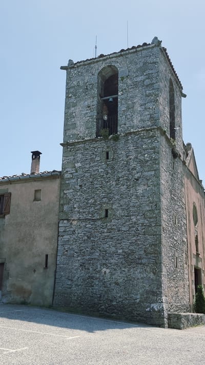 The image shows a close-up, low-angle view of a stone bell tower and an adjacent building in the village of Susqueda, Spain. The bell tower, made of rough-hewn gray stones, features a large arched opening containing a bell and a metal grate. Above the bell tower, the sky is clear and pale blue. To the left of the bell tower is a lighter-colored, stucco-clad building with a red-tiled roof and a visible chimney. The foreground is a paved surface, possibly a courtyard or street, with white lines marking an area. The overall scene depicts an old architectural structure under a bright, sunny sky.