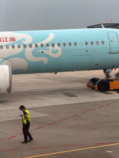 The image depicts the side of an aircraft with a light blue and white livery featuring wave-like designs, along with text in Chinese characters. An aircraft tug is partially visible near the nose of the plane. A ground worker, wearing a high-visibility yellow vest and dark trousers, stands on the concrete tarmac next to the aircraft. Red and yellow lines mark the tarmac surface, potentially indicating designated pathways or zones for aircraft operations. The setting appears to be an airport apron, most likely located in Baiyun District, China, as evidenced by the aircraft's livery which might indicate it is a Zhejiang Loong Airlines aircraft, a Chinese carrier. The sky is overcast, suggesting possible inclement weather.