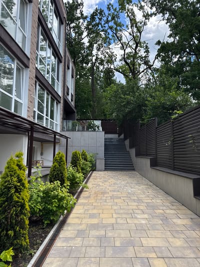 An exterior, eye-level, vertical shot shows a paved walkway leading towards a modern building and an outdoor staircase. The building is multi-story with a brick facade and large, reflective windows. A covered walkway extends from the building's ground floor, with a metal awning supported by dark brown posts. A row of manicured evergreen bushes and smaller leafy plants is situated along the left side of the paved walkway, separated by a metal grate drainage system. The walkway itself is constructed of interlocking stone pavers in shades of tan and grey. To the right, a dark brown slatted fence runs along a concrete retaining wall, partially obscured by trees. The background features lush green trees with a cloudy sky visible through their branches. The scene depicts a residential or commercial property, likely in Irpin, Ukraine, given the context. There are no people or animals visible in the image, and no apparent damage or signs of conflict are evident.