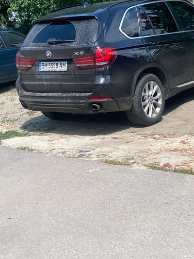 A black BMW X5 is parked on a dusty patch of ground, possibly a parking lot or roadside in Trostianets, Ukraine. The vehicle is oriented with its rear facing the camera. The license plate reads "BM 5558 AM." The car is slightly dirty, with dust accumulated on its surfaces. In the background, there are green trees and foliage, suggesting an outdoor setting. The foreground consists of a paved surface, likely a road or sidewalk, with some scattered debris. No people are visible in the image.