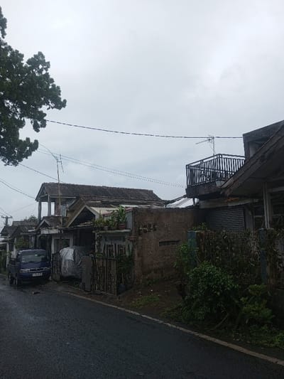 A row of houses lines a narrow road under a gray, overcast sky. The houses vary in design, with some featuring balconies and others appearing more modest. A dark blue pickup truck is parked along the road, near a car covered by a tarp. The houses are close together, with fences and vegetation separating them. Power lines stretch across the sky above the rooftops. The wet asphalt suggests recent rain.