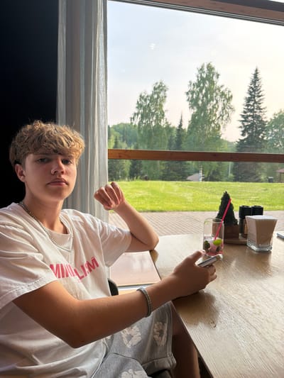 A young male with blond, curly hair is seated at a wooden table, looking directly at the camera. He is wearing a white t-shirt with pink text and grey shorts with a white heart pattern. A silver chain necklace adorns his neck, and a silver bracelet is on his left wrist. He holds a smartphone in his right hand.
On the table in front of him are a clear glass with a pink drink and mint leaves, a small decorative evergreen tree, salt and pepper shakers, and a container of napkins. The table is positioned near a large window that looks out onto a lush green lawn with trees in the background. The scene appears to be set in a cafe or restaurant in Staroabashevo, Russia. The overall lighting suggests it is daytime.
