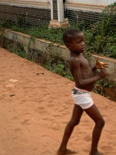 A young boy, likely of African descent, is captured standing on reddish-brown sand or dirt ground. He is shirtless and wears white briefs. The boy's right hand is raised to head level, forming a "peace sign" with two fingers extended, while his left arm is bent with the hand near his chest. He appears to be posing or playfully engaging.

In the background, behind the boy, there is a dense area of green foliage and overgrown bushes. Beyond this vegetation, a building with distinct architectural features is partially visible, including white window frames or pillars and dark brickwork. The ground beneath the boy shows subtle textures, possibly footprints or minor debris.

The lighting in the image is soft and even, suggesting either an overcast day or a time of day like late afternoon. The boy's expression is neutral to slightly playful. No visible text is present in the image. The scene is set in Aguleri, Nigeria. The photograph itself is oriented sideways, with the boy's vertical form appearing horizontal within the frame.