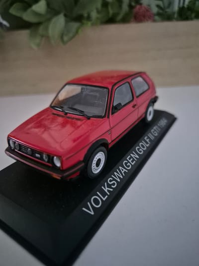 A red, die-cast model of a 1984 Volkswagen Golf GTI is displayed on a black base. The base has the text "VOLKSWAGEN GOLF II GTI 1984" printed in white letters. The model is positioned at an angle, with the front of the car facing the viewer's left. The background is blurred, showing a light-colored wooden surface and green foliage. There are no people visible in the image. The scene is indoors, with artificial lighting. There is no indication of the location being in Iskrets, Bulgaria.