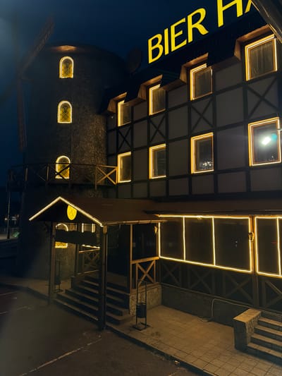 The image shows a building with a windmill tower, illuminated by warm yellow lights at night. The building has a traditional German-style facade with dark timber framing against a lighter exterior. There are multiple windows with yellow lighting outlining them. A large yellow sign spelling "BIER HA" is visible at the top of the building. The windmill tower has stone cladding and arched windows with similar yellow lighting. A wooden balcony or walkway extends from the windmill tower. An entrance area with a covered porch and wooden railings is also visible, illuminated by yellow lights. Steps lead up to the entrance. The ground in front of the building appears to be paved. In the distance, other buildings and lights are faintly visible. The overall scene suggests a restaurant or bar, likely a German-themed establishment. The presence of a "BIER HA" sign strongly indicates a "Bier Hall" or similar establishment. Based on the context provided that this image is from Ulan-Ude, Russia, this establishment is likely a local business catering to patrons seeking a particular atmosphere or cuisine.