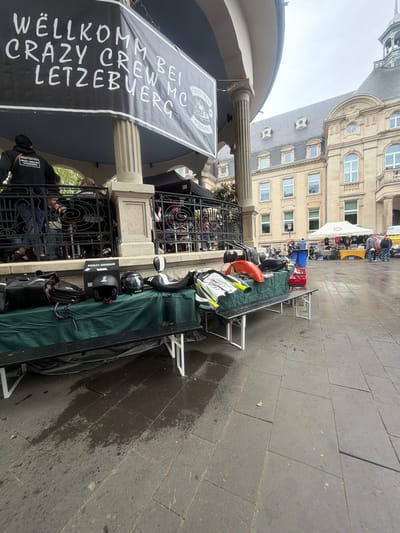 The image shows an outdoor market stall in Dudelange, Luxembourg, with a banner that reads "WELLKOMM CRAZY CREW MC LETZEBUERG" (Welcome Crazy Crew MC Luxembourg). Several motorcycle helmets and accessories are displayed on tables, including an orange fender and a Harley-Davidson box. People are seen behind a wrought-iron fence, and a building with multiple windows and a white tent are visible in the background. The ground is paved with gray stone tiles, some of which appear wet.