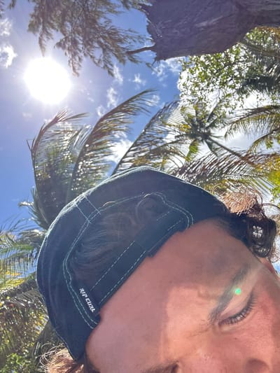 The image shows a low-angle view of a person wearing a dark blue Rip Curl cap with green stitching, positioned against a backdrop of palm trees and a partly cloudy blue sky. The sun is visible and bright, causing lens flare. The location appears to be outdoors, with tropical vegetation, likely in Sainte-Anne, France.