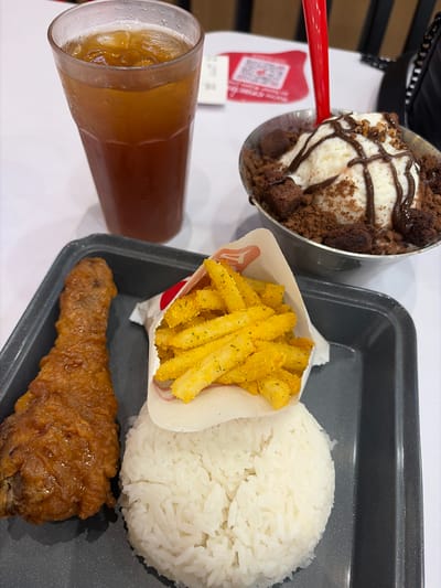 The image shows a meal laid out on a white table, with a black tray holding some of the food items. In the tray, there is a portion of white rice, a paper cone filled with golden french fries seasoned with herbs, and a piece of fried chicken. Next to the tray, a tall, clear glass contains a brown beverage with ice. To the left of the glass, a metallic bowl is filled with a dessert, likely ice cream or shaved ice, topped with chocolate syrup and crumbled chocolate cake or cookies. A red spoon is placed in the dessert bowl. A QR code is visible on a white paper in the background. The overall scene appears to be a casual meal at a restaurant or food establishment. The lighting suggests it is daytime.
