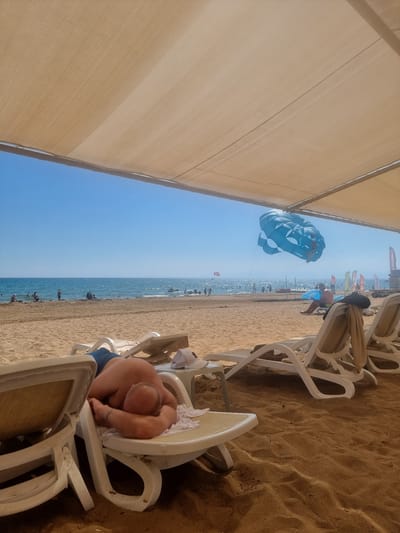 This media file captures a sunny day at a beach in Manavgat, Turkey. The scene is dominated by a large, tan canopy providing shade over a sandy area where white plastic lounge chairs are arranged. In the foreground, a person is lying down on a lounge chair, curled up and seemingly asleep, wearing blue shorts.
Further down the beach, the turquoise ocean stretches out to the horizon under a clear blue sky, suggesting fair weather. Several people are visible in the water, some appearing to be swimming or wading. A prominent blue and white parachute is in the air, indicating parasailing activities are taking place.
In the background, there are more lounge chairs and beachgoers. Some flags and what appear to be signs or banners are visible along the beachfront, adding to the lively atmosphere of a resort or tourist area. The time of day appears to be daytime, likely late morning or early afternoon given the bright sunlight and long shadows cast by the canopy and chairs. The overall emotion conveyed is one of relaxation and enjoyment of a beach vacation.