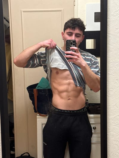 A shirtless man with abs poses in a mirror. He wears black sweatpants and a striped shirt partially pulled up. He holds a phone with a topographical map case, likely taking a selfie. The mirror reflects a partially visible room, including a door and what appears to be a portion of a dresser or cabinet. The photo is taken indoors with artificial lighting.