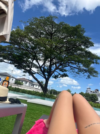 In Ratsada, Thailand, a person is reclining on a lounge chair in a sunny outdoor setting. The foreground features the person's legs, with a small tattoo visible on the right thigh. On a table beside the lounge chair are a straw hat with a black ribbon, a pair of sunglasses, and a bottle of "Australian Gold" sunscreen. A bright pink towel is draped over the lounge chair. In the mid-ground, a swimming pool with white lounge chairs is visible. Behind the pool, there is a well-maintained lawn and a fence. A large, leafy tree dominates the background, with its branches extending across the frame. The sky is blue with scattered white clouds. Residential buildings and some electrical poles are visible in the distance.