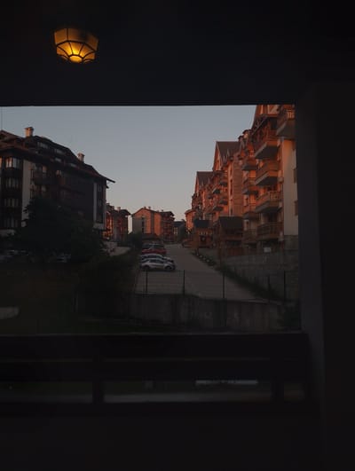 The image depicts a street scene in a mountainous resort town, likely Bansko, Bulgaria, during the golden hour. The warm, soft light of the setting sun casts a golden glow on the multi-story buildings that line a gently sloping road. These buildings appear to be hotels or apartment complexes, with balconies and traditional architectural elements, suggesting a European alpine setting.
Several cars are parked along the road and in a lot on the right side of the image, indicating it's a populated area. The vegetation, including trees, appears lush, suggesting a warmer season. The sky is clear and gradually transitioning from a pale blue to a darker hue near the top of the frame, where a decorative light fixture with a warm, yellow glow is visible.
The foreground is dominated by dark, shadowy elements, possibly a balcony or interior space from which the photo is taken, creating a framed view of the exterior scene. This framing adds a sense of depth and perspective. The overall mood is serene and peaceful, bathed in the tranquil light of dusk. There is no visible text in the image.