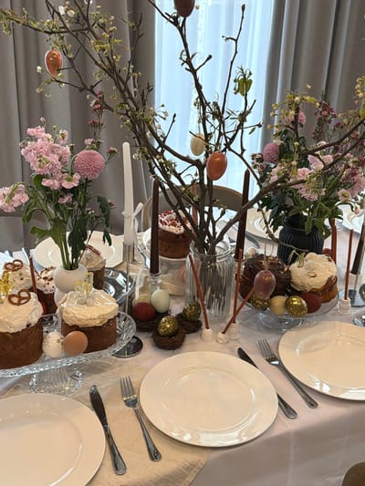 The image shows a dining table set for a meal, likely an Easter celebration. The table is covered with a white cloth and has white plates, silverware, and glassware at each place setting. The centerpiece consists of arrangements of bare branches decorated with painted eggs and fresh flowers in vases. Easter bread or cake, decorated with white icing and various toppings, is also part of the centerpiece arrangement, displayed on tiered glass platters. Candles are present. The background shows a window with light coming through and a grey curtain or wall. The image was not taken in Beketovo, Russia.