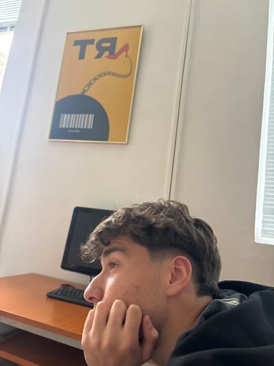 The image displays a young male in an indoor setting, likely a room in Troyan, Bulgaria, during the daytime. The individual, seen from a high angle and in profile, has short, curly dark hair and fair skin with some minor acne. He is wearing a dark hooded sweatshirt and is resting his left hand against his chin or cheek, looking downwards with a contemplative or focused expression.
Mounted on a light-colored wall behind him is a rectangular framed artwork or poster with a golden border. The poster features a vibrant yellow background. On the left, stylized text appears to be the Cyrillic letters "ТЯ" (which translates to "she"). A golden chain extends from the letter "Я" towards a large dark grey or black circular shape on the right side of the poster. Inside this circle, there is a white barcode pattern, and below it, faint, blurry white text that could be "HECTORY" or "НЕСТОРY".
Further to the right and slightly behind the person's head, a black electronic device, possibly a tablet or a small monitor, is visible. On a light brown wooden surface, presumably a desk, a black remote control or small keyboard is placed. The room is brightly lit, indicating natural light entering from a window or light source, partially visible with blinds in the upper and lower left corners. The scene suggests a calm and quiet atmosphere, with the individual appearing to be in thought or focused on something out of frame.