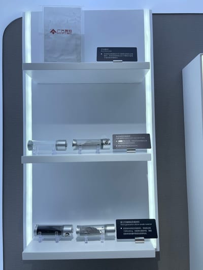Three white shelves with white trim, mounted to a white wall, display small, cylindrical, clear containers with silver caps, holding samples of silicon anode material. Each shelf includes a small black sign describing the product and its features. The shelves are lit with white lights. The logo for HYFTEC is displayed on a white paper on the top shelf. The location is Hangzhou, China at the coordinates (latitude: 30.272, longitude: 120.416).