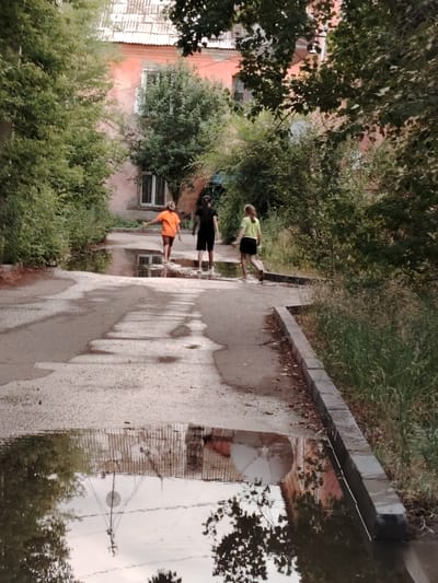 Three children are walking through puddles on a paved road. The child in the middle wears an orange t-shirt and shorts, and is barefoot. The child to their left wears a black t-shirt and shorts, and is also barefoot. The child to their right wears a yellow t-shirt and black shorts, and is also barefoot. They are walking away from the viewer, towards a building in the background. The building is pink and has several windows. There are trees and foliage on either side of the road, and the sky is reflected in the puddles. The scene appears to be in a residential area. The time of day is likely late afternoon or early evening, given the lighting and the reflections in the water. The weather seems to be overcast or recently rained, with large puddles on the road. This appears to be a snapshot of everyday life in Pavlodar, Kazakhstan, with children enjoying a moment of playful freedom after some rain. There is no visible text in the image.