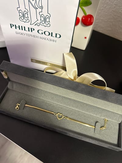 The image shows a gold bracelet with a heart charm in a gray velvet box. The box is partially open, and a cream-colored satin ribbon is tied around it. To the left of the box is a white gift bag or box with "PHILIP GOLD" printed in dark green and Cyrillic text below it. In the background, there is a white ceramic dish with two red cherry decorations. The scene appears to be indoors, on a dark, possibly black, surface. The location is not discernible from the image and is not relevant.