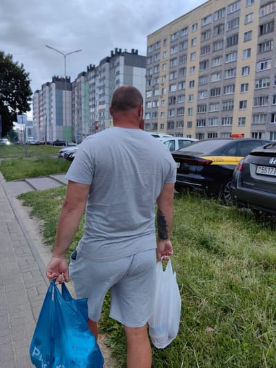 A man with a tattoo on his left arm, seen from behind, walks along a sidewalk. He is wearing a grey t-shirt and grey shorts and carries two plastic bags, one blue and one white, filled with groceries. The blue bag has visible text that appears to be "Pharia".
The man is walking in Fanipal, Belarus, as indicated by the location context. In the background, there are several tall, modern apartment buildings with light-colored facades and many windows. A street light is visible between the buildings. Cars are parked on the side of the road, including a black car with a yellow stripe and a license plate that reads "5355". Another grey car has a license plate ending in "5517". The grass on the side of the road is tall and overgrown, with some small pink flowers scattered within it. The sky is overcast, suggesting it might be late afternoon or evening, or perhaps a cloudy day. The overall scene depicts a residential area in Belarus.