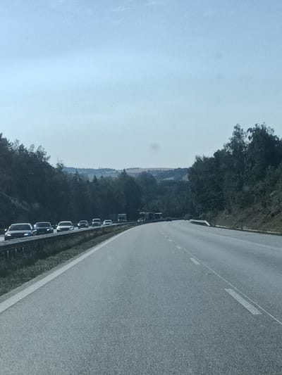 The media file shows a highway in a rural setting in Drahňovice, Czechia. The scene is dominated by a paved road stretching into the distance, with a lane of traffic moving away from the viewer. On the left side of the road, a line of cars is visible, traveling in the same direction. Beyond the cars, a dense forest lines the highway, with rolling hills and trees in the background. The sky is overcast, suggesting it is either early morning or late afternoon. There are no discernible people visible in the media file, and the focus is on the road and the surrounding natural landscape. The overall mood is one of a typical highway journey, with the natural scenery providing a backdrop to the movement of vehicles.