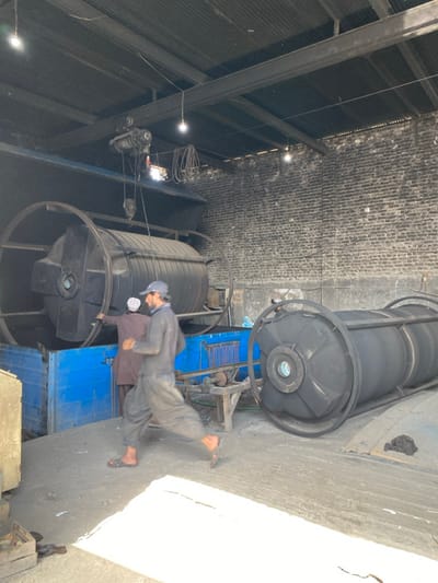 The scene depicts the interior of an industrial workshop or factory in Daman, Afghanistan. The area features a corrugated metal roof supported by dark I-beams, from which several bare LED light bulbs and an overhead crane system with a hoist mechanism are suspended. The walls are constructed from dark, exposed brick, with a lighter concrete base. The floor is an uneven concrete or dirt surface, illuminated by patches of natural light entering from an unseen opening.
Two large, dark grey, cylindrical rotational molding machines are present. One machine is horizontally positioned within a prominent blue rectangular metal housing or frame located in the mid-ground left, appearing to be in operation or being tended to. The second identical machine lies horizontally on the floor to the right, alongside a crude wooden table or support structure.
Two men are visible. In the foreground, a man with a dark baseball cap and a beard, wearing dark long-sleeved workwear and sandals, is captured in motion, blurred as he walks or runs from left to right across the frame. In the background, partially obscured by the moving man and the machinery, a second man wearing a light-colored cap and light brown traditional attire stands next to the active blue housing, with his arm extended towards the rotating machine. Various industrial cables and wires are visible throughout the overhead structure.