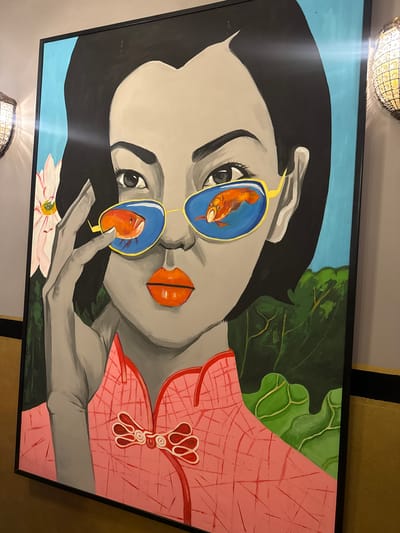 The image shows a close-up of a large painting featuring a stylized portrait of a woman. The woman has dark hair, grey skin, and large orange lips. She is wearing mirrored sunglasses with gold frames, and the lenses reflect two orange goldfish. Her right hand is raised, with fingers near her sunglasses. She is wearing a pink garment with a traditional Chinese knot closure. Behind her, to the left, is a white and pink flower, possibly a lotus. To the right, there are large, dark green leaves. The painting is framed in black and hangs on a wall. The wall is painted in muted tones, with a textured yellow stripe visible at the bottom and right edges of the frame. Two ornate light fixtures, one on the left and one on the right of the painting, illuminate the scene with warm light. The lighting casts a noticeable glare across the painting, particularly on the upper right side. The overall impression is of a modern art piece displayed in an interior setting. This painting is located in Barvikha, Russia.
