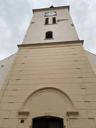 The image shows a low-angle, full-frame shot of the tower of the Church of Saint Nicholas in Bílina, Czechia. The tower has a white facade with some visible wear and tear, particularly on the upper section where plaster is missing. A clock face with Roman numerals is visible near the top of the tower. Below the clock, there are two arched windows with dark shutters. The lower section of the tower is covered in a beige, textured material, divided into horizontal bands with a staggered pattern. An arched entryway with a dark wooden door is visible at the bottom of the tower. The sky above is overcast and grey. The image focuses solely on the architecture of the church tower.
