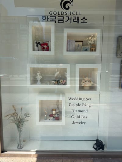 The image shows a storefront window display of a jewelry store. The store name, "GOLDSHELL," is visible at the top of the display, along with Korean text that translates to "Korea Gold Exchange." The display features several illuminated square frames showcasing various items.

In the top left frame, there is a gift-wrapped box with a black ribbon and a small teddy bear made of pink flowers. The top right frame contains two books, a framed picture, a small wooden chair, and a decorative floral arrangement.

The middle left frame holds a white bust and a bouquet of pink and peach flowers. The middle right frame displays a cylindrical vase filled with pale pink flowers and reeds.

The bottom left of the display has a tall clear vase filled with dried grass-like reeds. The bottom right frame contains a small decorative bucket, a framed floral picture, and what appears to be a small bottle with reeds.

Below the display frames, a list of items is printed: "Wedding Set," "Couple Ring," "Diamond," "Gold Bar," and "Jewelry." At the bottom right corner of the display, a small black vintage-style camera is placed. The reflections on the glass suggest this is an outdoor setting, likely on a street. The tiled flooring is visible in the foreground.

This storefront is located on the street of a city. The visual cues, including the Korean text and the style of the storefront, suggest a location in South Korea. Based on the architectural and urban elements, it is plausible this is in Busan, South Korea.