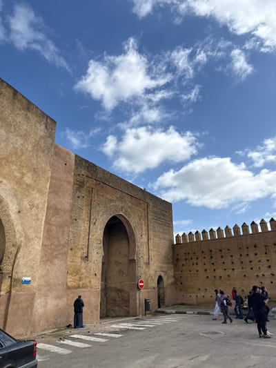 A daytime street scene in Mechouar Fes Jdid, Morocco, depicts a historic city wall and gateway under a partially cloudy blue sky. The ancient reddish-brown wall, constructed of varying stone, features multiple large arched entrances. On the left, a person wearing a dark djellaba and a black puffer jacket is performing prayer on a blue mat, facing an arched alcove in the wall. A black sedan is partially visible in the bottom-left corner. To the right of the praying individual, a red circular "No Entry" sign is mounted on a pole, with a black trash receptacle beneath it. Further to the right, the wall extends with a crenelated top and a section punctuated by numerous small circular indentations. A group of approximately ten individuals, dressed in a mix of traditional and modern attire, gathers on the right side of the paved street near a vendor's display of produce. One individual in black clothing is observed photographing the scene with a smartphone. White crosswalk stripes are visible on the asphalt road in the foreground.