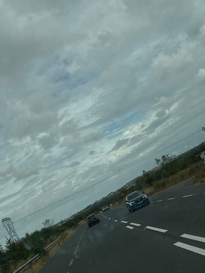 The image depicts a section of a road, likely a highway or a major route, in the city of New Ross Municipal District, Ireland. The scene is captured from a moving vehicle, as evidenced by the perspective and motion blur. The road is asphalt and marked with white dashed lines indicating lanes. Several cars are visible, with one dark-colored hatchback in the foreground on the right lane, its headlights illuminated. Other vehicles are further down the road in the distance.
The environment surrounding the road is rural, with green vegetation, dry grasses, and scattered trees. In the background, rolling hills and a cluster of buildings suggest a semi-rural or suburban setting. A prominent industrial structure, possibly a pylon or a power line tower, is visible on the left side of the image, adding an element of infrastructure to the landscape.
The sky is overcast with dense, grey clouds, indicating a possibly inclement weather day. The overall lighting is dim, consistent with a cloudy or late afternoon sky. The image is slightly tilted, suggesting the photograph was taken at an angle. No people are clearly discernible within the frame, and the focus is on the road and its surroundings.