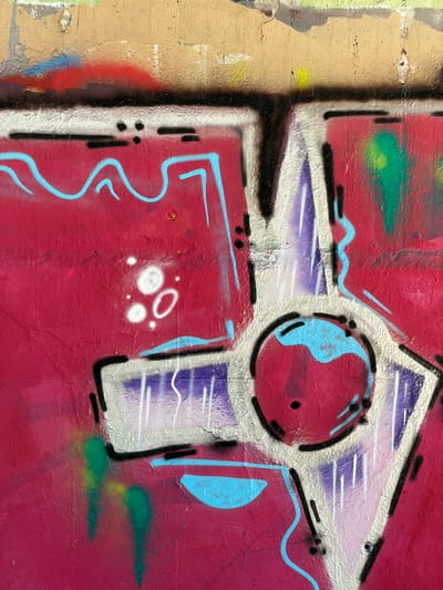 The image is a close-up, vertical shot of a graffiti-covered wall in l'Hospitalet de Llobregat, Spain. The graffiti features vibrant colors, primarily a deep magenta background with accents of bright blue, purple, white, and green spray paint. Abstract geometric shapes and fluid lines dominate the composition. Some areas of the wall appear weathered and textured, with remnants of older paint or plaster visible. There are no people or discernible actions depicted; the focus is solely on the colorful, abstract street art.