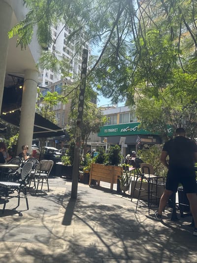 The image displays an outdoor street scene, likely a commercial or cafe district, under bright daylight. Lush green trees with delicate leaves frame the upper portion of the frame, casting dappled shadows across the paved sidewalk and various objects below.

In the foreground, several outdoor cafe tables and chairs, some featuring black and white woven patterns, are arranged on the sidewalk. A large concrete column partially obscures a person seated at one of these tables on the left. Further to the right, a person dressed in a black t-shirt and shorts stands near another cafe table, their back mostly to the viewer. A wooden planter box is also visible.

Across the street, buildings line the background. One prominent building features a green awning with visible text. The English words "DELI MARKET" are clearly displayed in white capital letters. To the left of "DELI MARKET," Hebrew script is visible, which translates to "Ma'adaniya" (delicatessen), providing a strong geographical cue. A distinctive street pole adorned with white circular patterns and what appear to be small stickers or posters stands upright in the middle ground. Further down the street, additional buildings and glimpses of other people and vehicles can be seen, suggesting a bustling urban environment.

The strong, distinct shadows indicate a sunny day, likely mid-morning to early afternoon. The scene conveys a casual, relaxed atmosphere, typical of people enjoying an outdoor cafe setting. The visible Hebrew text and architectural style align with the given location context of Givatayim, Israel.