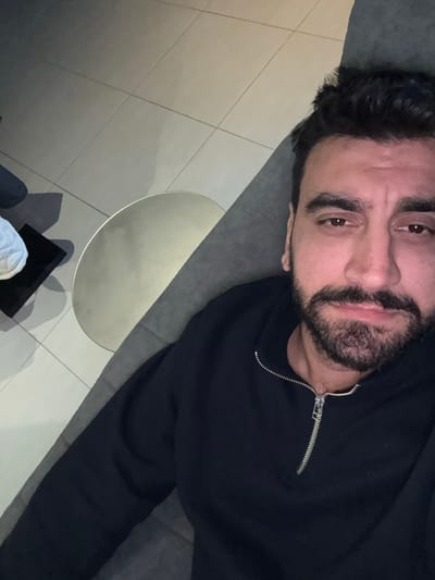 An adult male with dark hair, dark full beard, and brown eyes is depicted in an indoor setting in Mesa Geitonia, Cyprus. He is positioned reclining on a grey upholstered surface, likely a sofa, looking directly towards the camera with a neutral expression. He wears a dark, possibly black or navy, long-sleeved garment with a silver half-zip collar. To his lower left, a round, metallic silver-colored table is visible. Beyond the table, a light-colored object, possibly a shoe or cushion, rests on a dark rectangular object. The floor consists of light-colored square tiles with visible grout lines. The lighting suggests an artificial indoor light source.