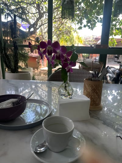 The scene is a table setting at a restaurant in Celuk, Indonesia. The table is marble and holds a plate, a cup, and a saucer. A silverware holder containing utensils and a container holding paper napkins are also on the table. A vase containing purple orchids provides a floral accent. Outside the window, lush tropical foliage and glimpses of buildings confirm the location. The presence of dining ware suggests a meal setting. No people are visible, focusing the scene solely on the arranged items. A glimpse of a scooter can be seen through the window.