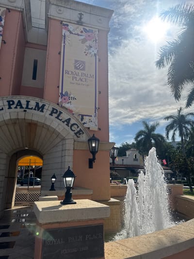 The image depicts the entrance of "Royal Palm Place" in Boca Raton, Florida, United States of America. The architectural style is Mediterranean Revival, characterized by its stucco exterior, arched entryway, and decorative banners. The sun shines brightly, casting a warm glow on the scene, with palm trees visible in the background. A multi-tiered fountain stands in the foreground, its water jets creating a dynamic visual element. The name "Royal Palm Place" is prominently displayed on the arched entrance and on a bronze plaque at the base of the fountain, which also indicates the founding year of 1962. The overall impression is one of a well-maintained, upscale commercial or retail space.