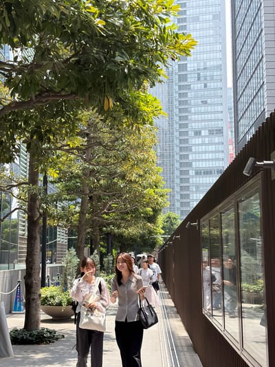 In Chiyoda, Japan, two young women walk side-by-side on a sun-drenched sidewalk adjacent to a building with a corrugated brown facade and large reflective windows. The woman on the left, wearing a light-colored blouse and dark pants, carries a white tote bag adorned with cartoon characters and a mobile phone. She has dark hair and is smiling while touching her mouth. The woman on the right, with brown hair, wears a gray collared shirt and a black skirt, and carries a black handbag. She is also smiling and looking towards the left. Behind them, several other people are visible, some walking and some reflected in the windows. Trees with lush green foliage line the walkway, with modern, glass-fronted skyscrapers towering in the background. The scene appears to be a bustling urban environment on a clear day, possibly a commercial or business district.