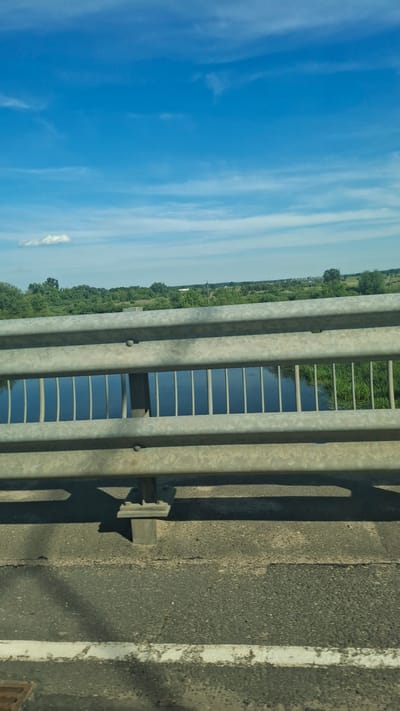 The image depicts a view from a bridge overlooking a body of water and a treelined landscape. A metal guardrail runs horizontally across the foreground, with a portion of the bridge's asphalt surface visible below it. The water appears calm, reflecting the blue sky and clouds above. Green vegetation lines the far bank of the water, with trees forming a dense backdrop. In the distance, hints of infrastructure, possibly power lines or a distant structure, can be discerned against the horizon. The sky is a vibrant blue, with scattered white clouds. The scene is devoid of human presence. The image appears to have been taken in Stowbtsy, Belarus, likely during daytime given the bright sunlight.