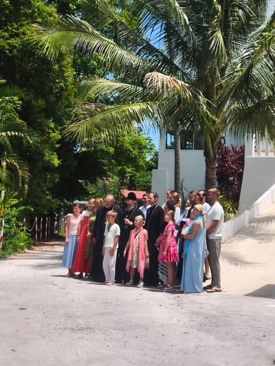 In Baan Hua Thanon, Thailand, a group of approximately twelve people, including adults and children, are gathered on a paved outdoor area, possibly a walkway or driveway, in front of a white building with a sloped roof. The group is positioned on the left side of the frame, with the building and lush green foliage, including palm trees, forming the background. Several individuals in the group are dressed in what appear to be religious or formal attire, with at least two men wearing black robes and head coverings, suggesting a religious ceremony or gathering. The people are looking in various directions, with some posing for a photograph. The environment is sunny, with shadows cast on the ground, indicating a bright day. The building appears modern, with white railings visible on a raised section.