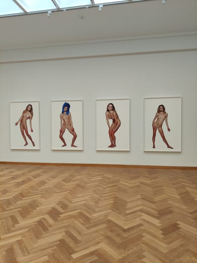 The image shows a gallery or exhibition space with four framed photographs displayed vertically on a white wall. The photographs feature nude female figures posed in dynamic, almost floating positions against a plain white background. The figures are rendered with a soft, almost ethereal lighting that accentuates their forms. One photograph features a woman with bright blue hair, while the others depict women with natural hair colors. The framing is simple and white, allowing the artwork to be the central focus.
To the right of the wall with the artwork, a polished wooden floor with a herringbone parquet pattern is visible. The lighting in the space appears to be artificial, consistent with an indoor gallery setting, and there are no external weather cues. The overall atmosphere is one of quiet contemplation, typical of an art exhibition. The setting suggests an indoor exhibition space, likely a museum or art gallery. No visible text is present in the image. The context provided indicates the location is The Hague, Netherlands.