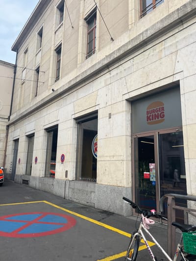 The image captures a street-level view of a multi-story building, likely in an urban setting. The building features a facade constructed from light-colored stone, with several rectangular windows visible on its upper floors.
The ground floor of the building hosts a commercial establishment, prominently a Burger King restaurant. Its classic oval logo, displaying "BURGER KING" in red text between two bun halves, is visible on a frosted glass window or door panel. Another, smaller circular Burger King logo is also discernible in a window further down the building's facade. The main entrance to the Burger King has glass doors adorned with multiple small, colorful stickers or signs.
Adjacent to the building is a paved area, possibly a sidewalk or part of a street. Distinct yellow lines are painted on the ground, and a large blue and red circular "no parking/stopping" symbol is painted directly onto the pavement. Additional smaller, circular "no parking" signs are affixed to the building's facade below the Burger King.
On the right side of the image, a light-colored bicycle is parked next to a metal railing. It has a green basket attached to the handlebars and a black seat. The brand "VILLIGER" might be visible on its frame. Overhead cables are also visible against the building and the sky. The time of day appears to be daytime, under what looks like an overcast sky, as no strong shadows are present. No people are visible in the scene.
