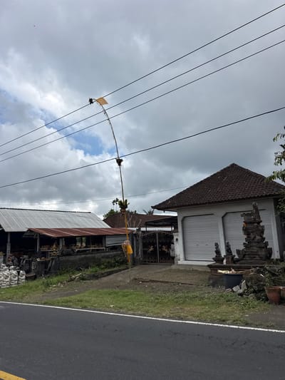 The image captures a roadside scene in the city of Susut, Indonesia, under a cloudy sky. A tall, slender pole with a yellow flag and some decorative elements stands prominently, reaching towards power lines that stretch across the frame. To the left, a long building with a corrugated metal roof, possibly a workshop or storefront, displays stacks of white bags and various items, some appearing to be sculptures or crafts, on shelves. In the center, a gate with ornate metalwork leads to a building with a tiled roof and two large, gray roller doors. To the right of this building, a stone shrine or sculpture stands in front of a gray wall, adorned with smaller stone figures. A dark bin and debris are present in front of the shrine. The foreground shows a paved road with a white line marking its edge and a yellow line on the far left. The environment appears rural or semi-urban, with vegetation and palm trees visible in the background.