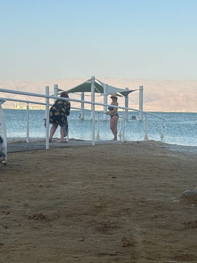 The image shows a beach scene with two individuals. The person in the foreground is facing away from the camera, wearing a dark dress with a floral pattern and standing on a wooden walkway. They are leaning on a white railing of a structure that appears to be a cabana or shelter, with a green canopy. Behind this person, another individual is visible, wearing a bikini and a hat. This person is facing the side and appears to be standing or posing near a similar structure.
The background features a calm body of water, likely the sea, with distant hills or mountains under a clear, pale sky. The lighting suggests it is either early morning or late afternoon, given the soft, diffused light. The beach itself is sandy and textured. The overall impression is one of a relaxed day at the beach, possibly a resort or public beach area given the presence of the shelters and railings. No visible text is present in the image. The scene is set in the Tamar Regional Council, Israel, hinting at a coastal or Dead Sea location.