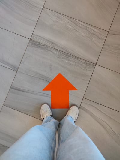 A person wearing light blue jeans and white sneakers stands on a tiled floor. An orange arrow is painted on the floor, pointing upwards. The tiles are large, rectangular, and have a light gray, marbled pattern. This image was taken in Khrabrovo, Russia.