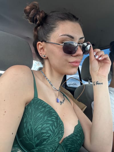An indoor, high-angle shot captures a woman seated in a car, likely in Belopoptsi, Bulgaria, during daytime. She is the primary subject, shown from the chest up, facing slightly away from the camera. Her dark hair is styled in a bun, and she wears multiple silver earrings on her left ear.
She is dressed in a dark green lace bra and a beaded necklace featuring a blue butterfly charm. Her left wrist displays a clear tattoo that reads "be brave," while another, less distinct tattoo is visible on her chest. Her nails are painted white. The woman is holding a pair of square-framed sunglasses, partially obscuring her face, with bright outdoor light reflecting in the lenses, suggesting a sunny day.
The car's interior, including a seat and a brown paper bag, is visible around her. Part of another person's arm and torso are faintly visible in the upper right corner of the frame. The woman appears relaxed, with her lips slightly parted. The scene conveys a casual moment inside a vehicle.