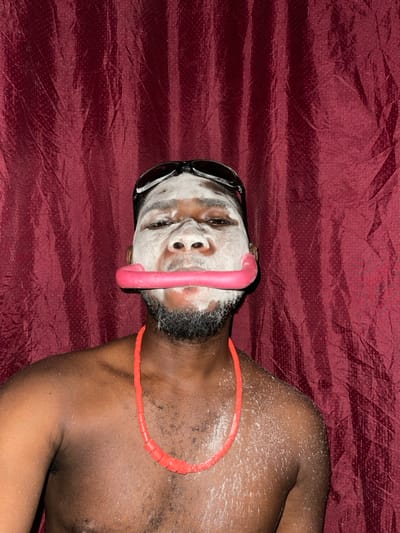 A man with sunglasses and a white face mask lies on a red, textured surface. A pink, hose-like object stretches across his face. He wears a segmented, orange necklace. His dark skin contrasts with the white mask and the surrounding red background.