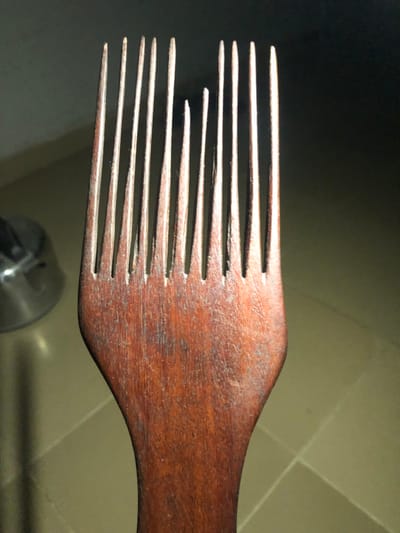A wooden hair pick with many tines is angled across the frame. The tines are not all the same length; some are broken or shorter than others. The pick shows wood grain detail and an overall reddish-brown tone. A blurry background suggests a light surface with hints of a metallic object. The lighting emphasizes the texture of the wood.
