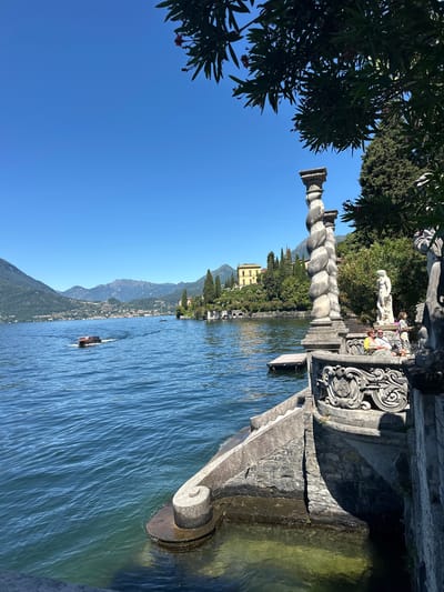 This is a full-frame, eye-level, outdoor, bright daylight shot of a serene lakeside scene, likely in Varenna, Italy, given the context of the image and common tourist destinations.
In the foreground, a stone balustrade, featuring intricate carvings and ornate scrollwork, forms the right edge of the frame. To the left of the balustrade are stone steps leading down to the water. A large, twisted stone column stands prominently behind the balustrade, partially obscured by lush green foliage overhead. To the right of the column, a white statue of a male figure stands on a pedestal, and two women are seated on a bench nearby. The water of a large lake, likely Lake Como, dominates the left side of the image. The water is a vibrant blue, with gentle ripples. A small boat with a red top is moving away from the camera, leaving a wake.
In the middle ground, across the lake, are the lush green hillsides dotted with buildings. A prominent yellow building is visible on the hillside. The landscape is characterized by rolling hills, lined with cypress trees and other greenery.
The background consists of a clear, bright blue sky. Distant mountains, with a haze, are visible on the horizon, framing the lake and the landscape on the opposite shore. The overall atmosphere is peaceful and picturesque, evoking a sense of tranquility and natural beauty.