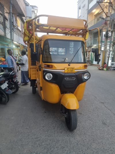 The image captures a yellow auto-rickshaw in Puttaparthi, India. The auto-rickshaw, bearing the license plate "AP39W J1385," is parked on a paved road. Two men are visible, one standing near the auto-rickshaw wearing a white shirt and dark trousers, and another man in a blue shirt examining a motorcycle. The street scene includes buildings, shops, and street vendors. The buildings have signs in Telugu and English, notably including a sign for "ROCS LAB & H". The overall environment suggests a typical street scene in Puttaparthi, with local businesses and transportation.