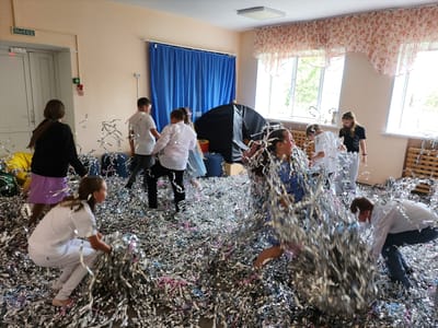 Location: Krasnogorskoye, Russia, interior of a room.
Scene: A group of children are actively playing with silver and pink confetti scattered across the floor. The children are dressed in a variety of clothing, including white shirts, dresses, and leggings. Some children are standing, walking, and bending over, gathering handfuls of confetti. The room appears to be a classroom or playroom, with a window covered by floral-patterned curtains. Blue curtains hang in the background. A sign with the word "ВЫХОД" (EXIT) in Russian is located near the door in the background. A small black tent is visible near the center.